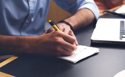 Man writing list next to laptop.